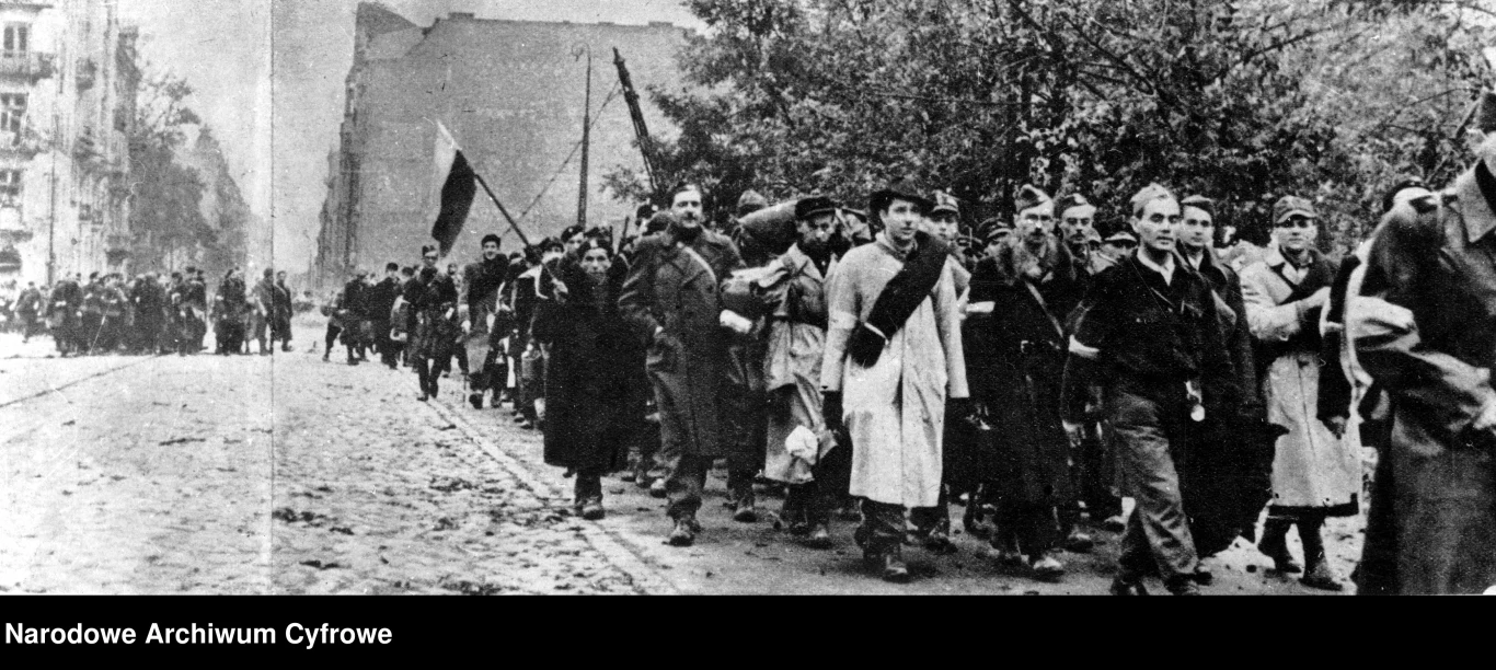 Kolumna pieszych ubranych w cywilne i wojskowe ubrania maszerująca miejską ulicą, część osób niesie flagę i torby, w tle zniszczone budynki oraz drzewa.