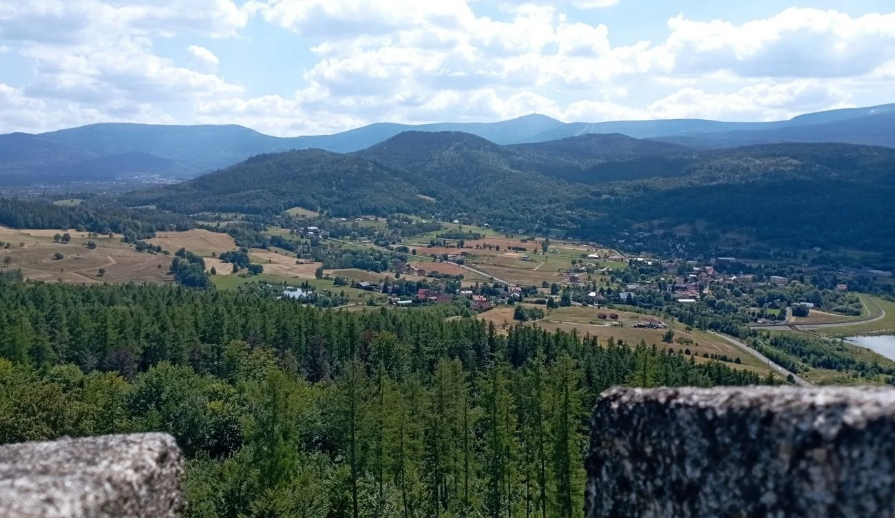 Zamek księcia Henryka - widok z wieży zamkowej. Rozległa panorama górskich wzgórz z licznymi połaciami lasu sosnowego, wiejskimi zabudowaniami i polami rozciągającymi się w dolinie, otoczona pasmem niebieskich gór na horyzoncie.