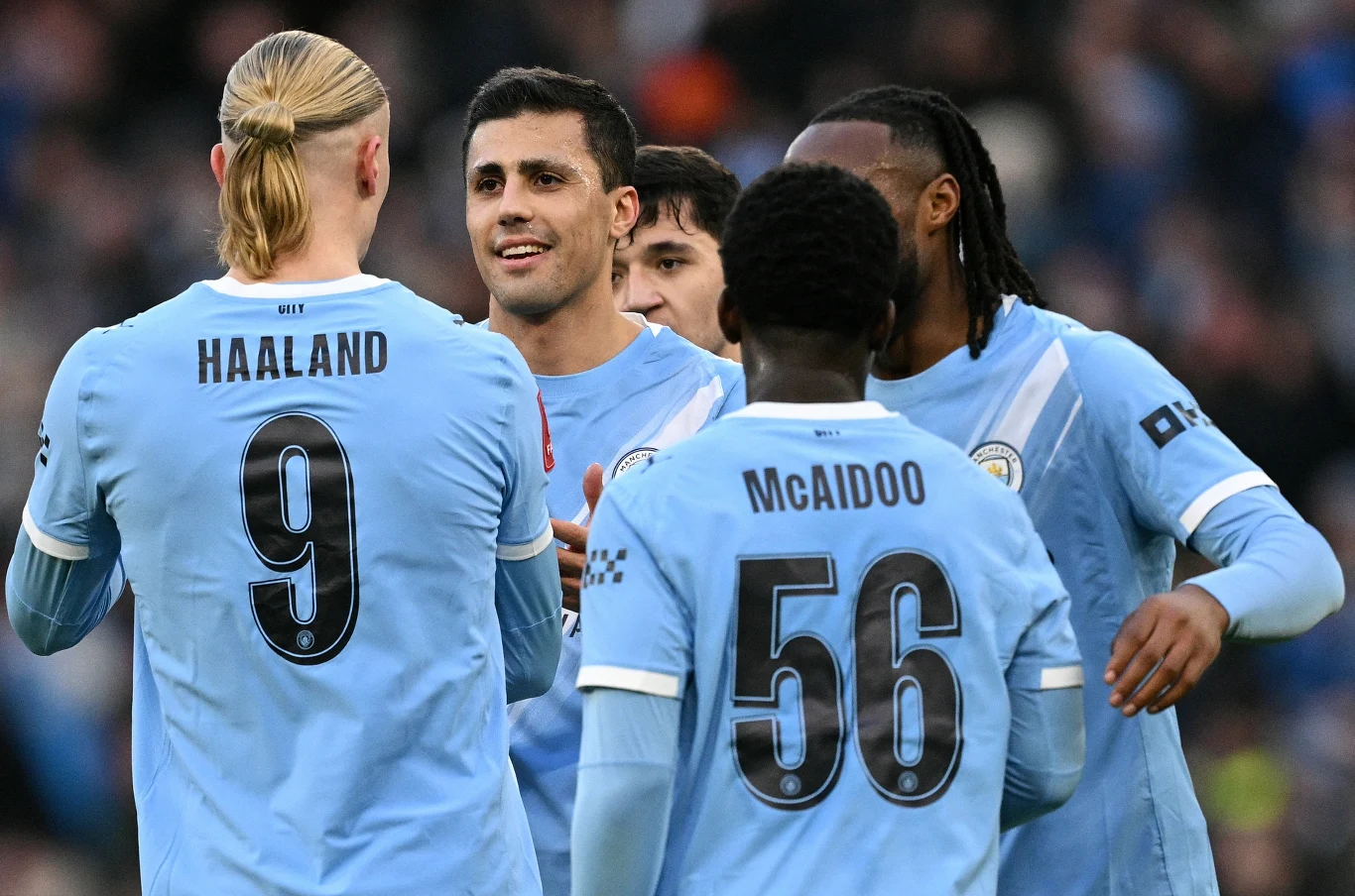 Pięciu piłkarzy Manchesteru City w błękitnych koszulkach, z których dwóch widocznych jest od tyłu z nazwiskami Haaland i McAidoo oraz numerami na plecach, celebruje wydarzenie podczas meczu.