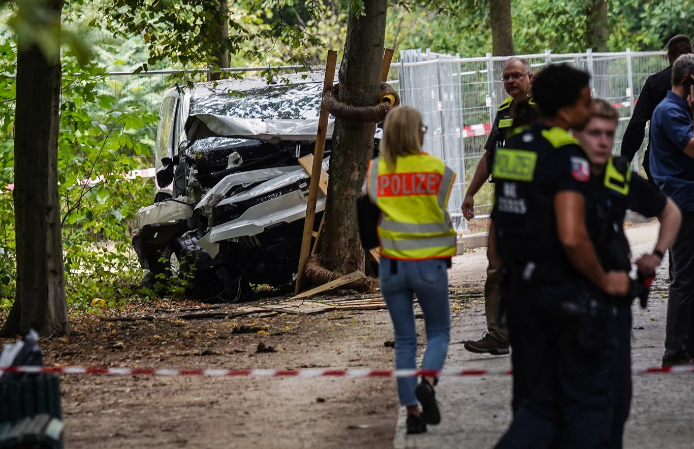 Niemieckie służby pracujące na miejscu tragedii. W ataku zginęła Polka Rozbity biały samochód wbity w drzewo, wokół miejsca wypadku stoją policjanci oraz śledczy, teren odgrodzony taśmą policyjną.
