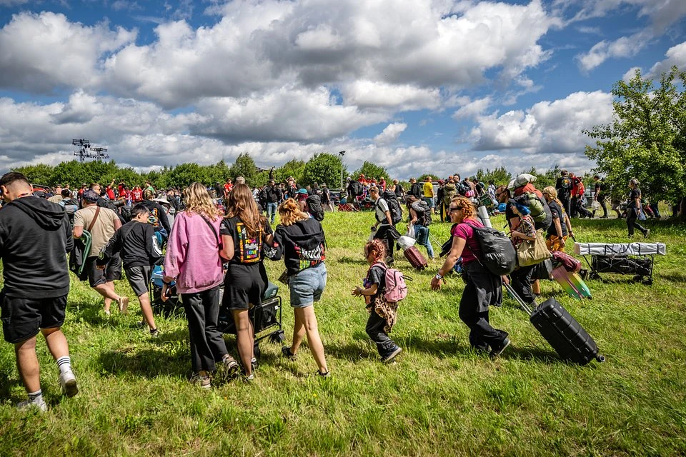 Ludzie przemierzający zieloną łąkę z plecakami i walizkami, zmierzający w stronę tłumu rozciągającego się na tle drzew i pochmurnego nieba.