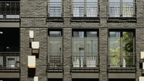 Ciemna ceglana elewacja budynku z dużymi oknami, metalowymi balustradami i nowoczesnymi, geometrycznymi lampami ściennymi.