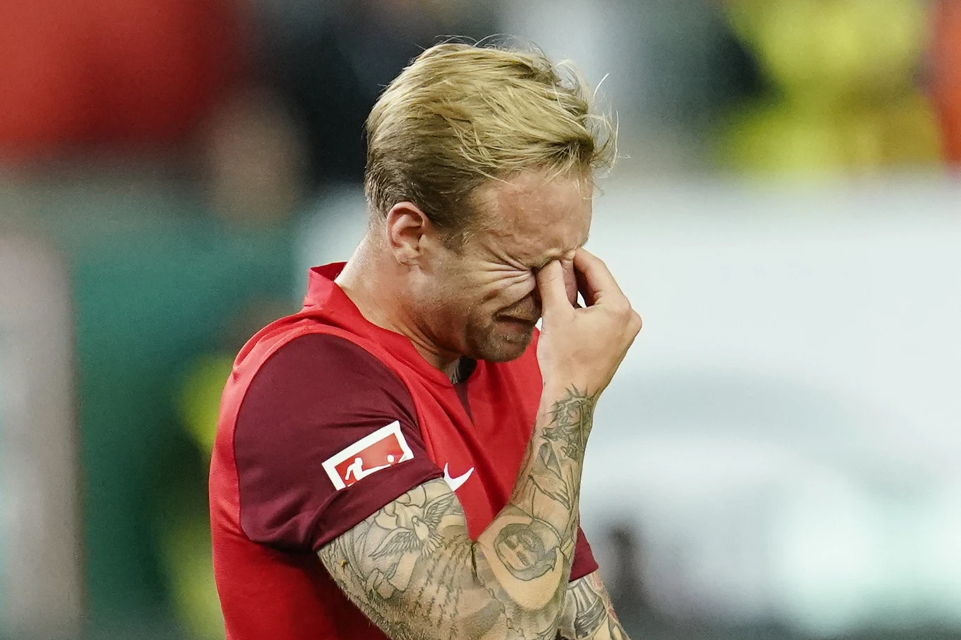Piłkarz w czerwonym stroju z tatuażami na ramieniu trzyma się za twarz, wyrażając silne emocje.