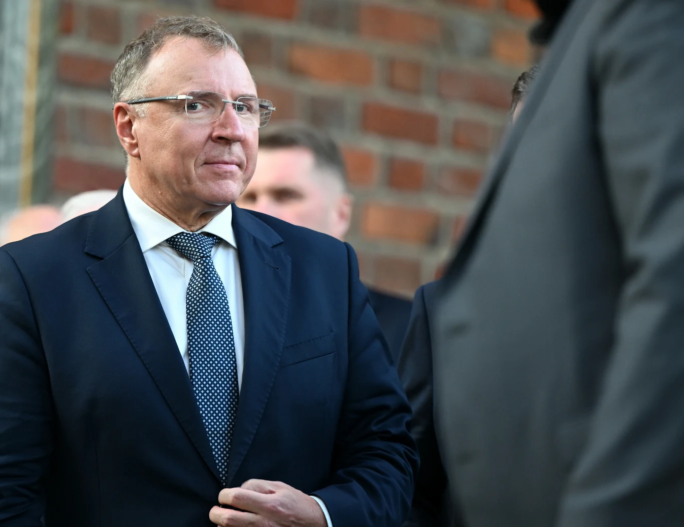 Jaka będzie przyszłość Jacka Kurskiego w PiS? Jacek Kurski w okularach i garniturze stoi na tle ceglanej ściany, wśród innych osób.