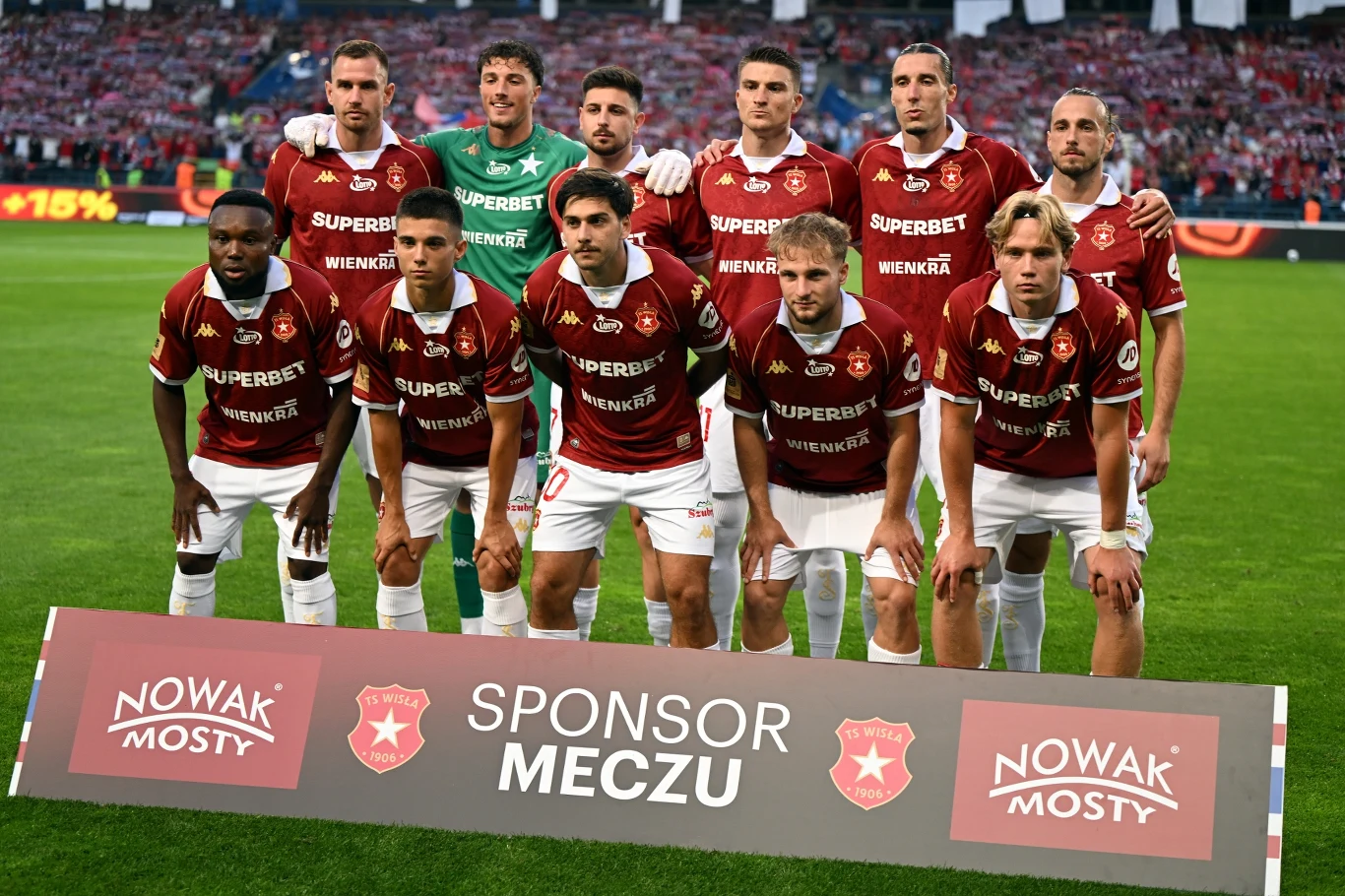 Piłkarze Wisły Kraków w sezonie 2026/2027 PKO BP Ekstraklasy Jedenastu piłkarzy w czerwono-białych strojach drużyny Wisły Kraków ustawionych w dwóch rzędach na murawie stadionu przed meczem, przed nimi tablica z logo sponsora i napisem 'Sponsor Meczu'.