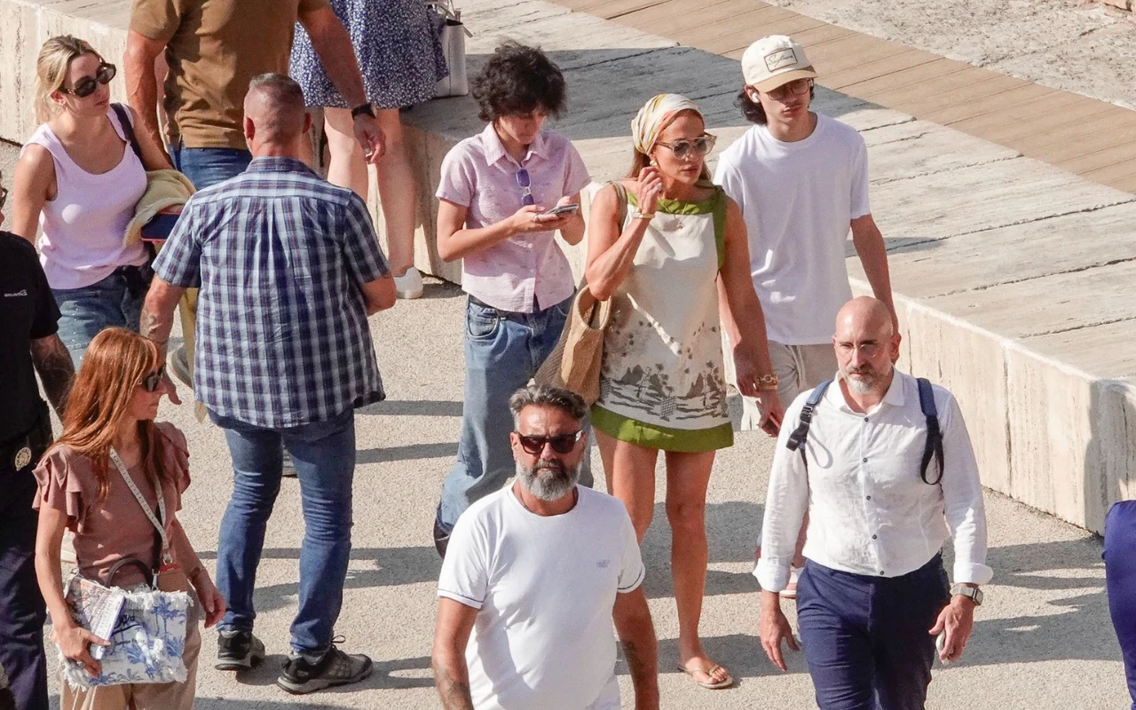 Grupa spacerujących ludzi, wśród których wyróżnia się kobieta w zielonej sukience i mężczyzna w białej koszulce z brodą; wszyscy ubrani lekko, sugerując letnią pogodę.