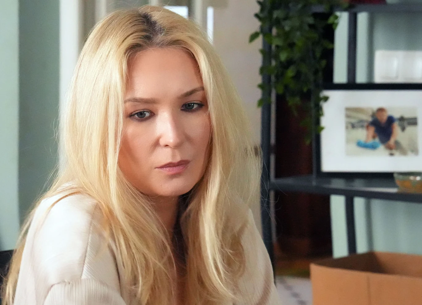 Kobieta o długich blond włosach siedzi zamyślona, w tle półka z ramką na zdjęcie i rośliną.
