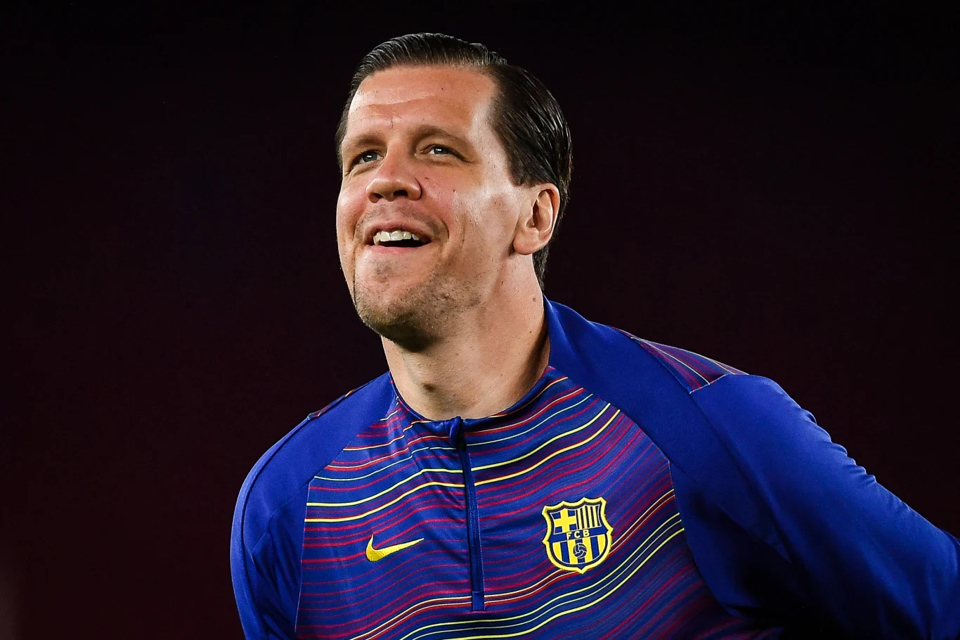 Wojciech Szczęsny Uśmiechnięty mężczyzna w sportowej bluzie FC Barcelony na ciemnym tle.