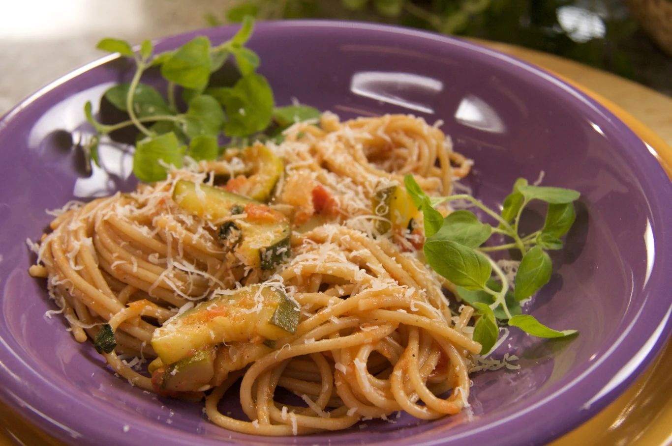 Makaron spaghetti z warzywami, startym serem i świeżą gałązką zielonych ziół na fioletowym talerzu.