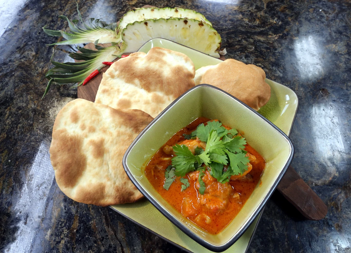 Miseczka z curry zwieńczonym świeżą kolendrą, obok trzy kawałki pieczonego chleba typu naan oraz plaster ananasa na talerzu.