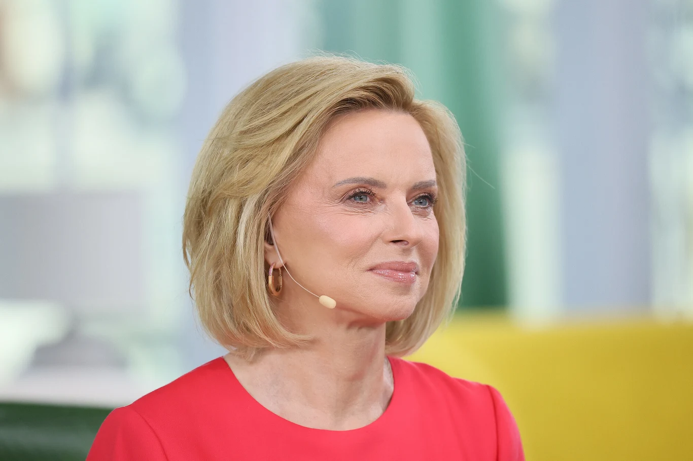 Uśmiechnięta kobieta z blond włosami do ramion, ubrana w różową sukienkę, z założonym przy uchu mikrofonem.