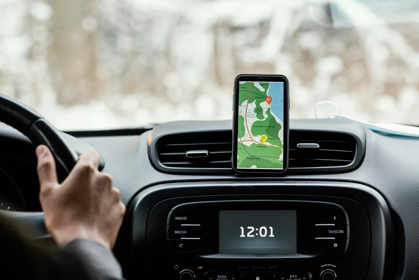 Kierowca trzymający kierownicę samochodu, na desce rozdzielczej zamontowany telefon z włączoną nawigacją GPS oraz wyświetlony zegar cyfrowy pokazujący godzinę 12:01.