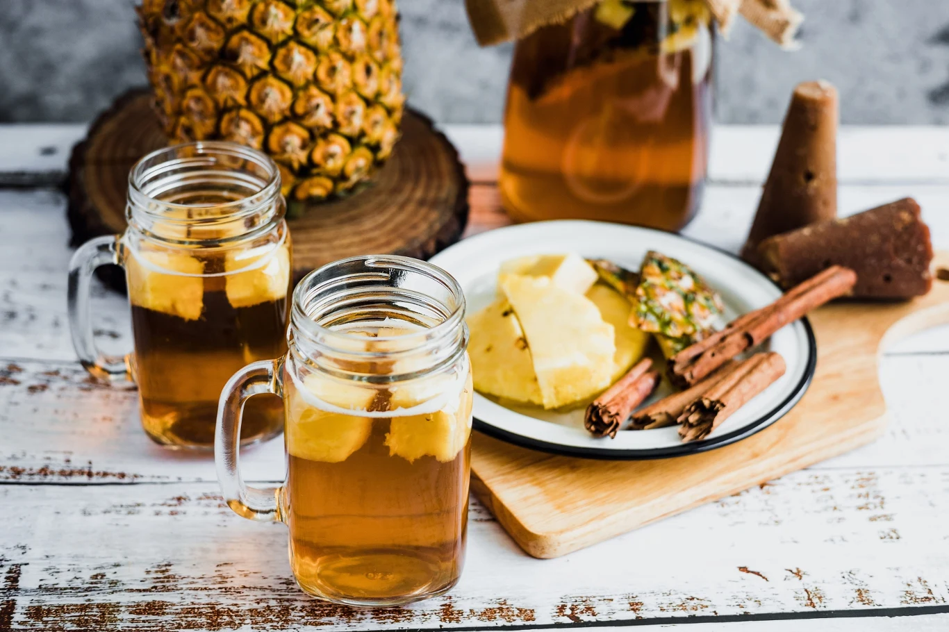 Szklanki z napojem zawierającym kawałki ananasa, obok talerz z plastrami ananasa, laskami cynamonu i kawałkiem słodkiego bloku, w tle świeży ananas oraz słoik z napojem na drewnianym podkładzie.