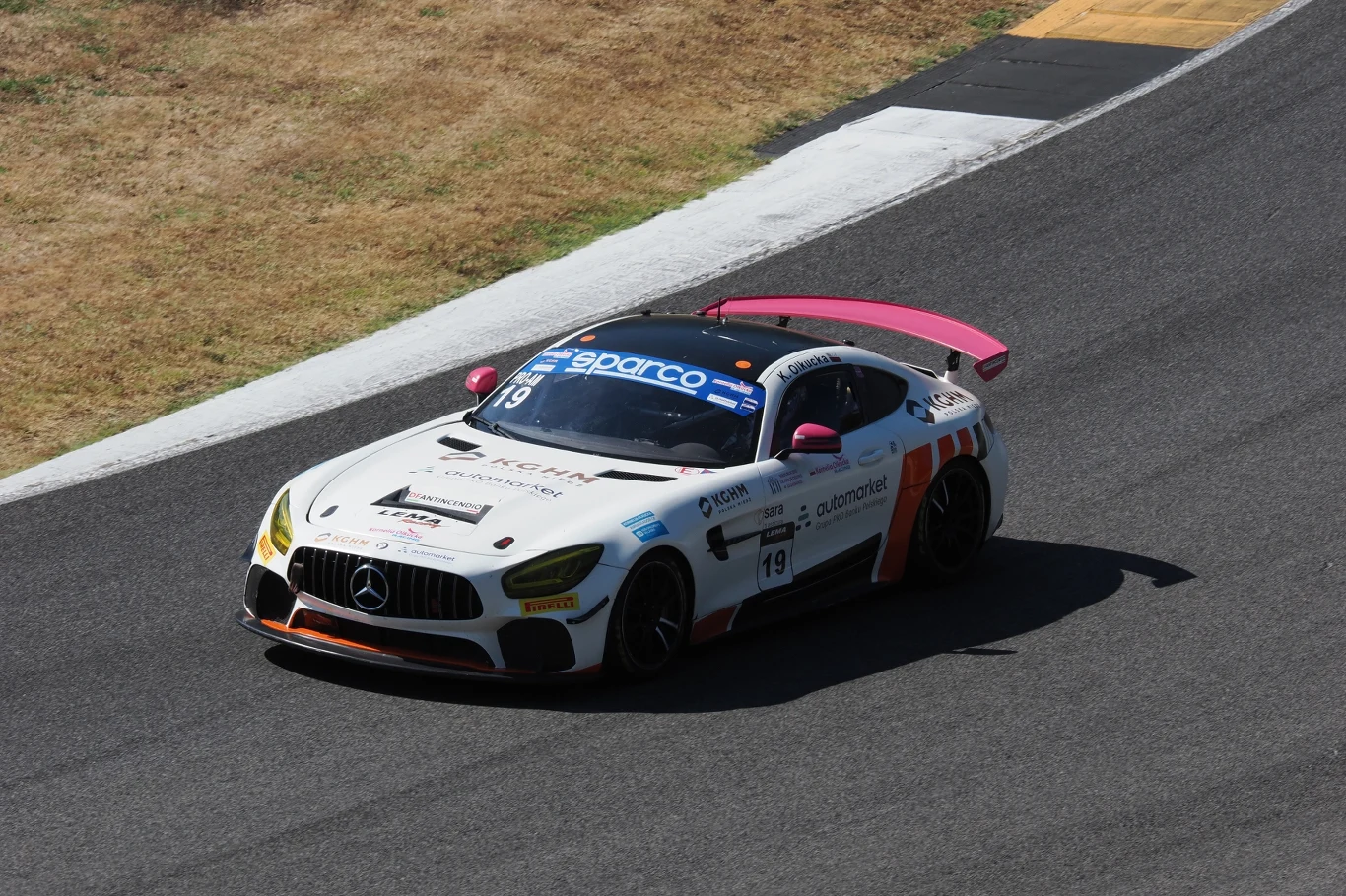 Wyścigowy samochód Mercedes AMG GT3 z numerem 19, biało-różowe barwy, dynamicznie pokonuje zakręt asfaltowego toru.