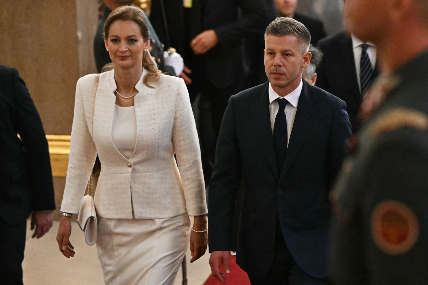 Elegancko ubrana kobieta w białym kostiumie i mężczyzna w ciemnym garniturze idą razem, otoczeni innymi ludźmi w formalnych strojach.
