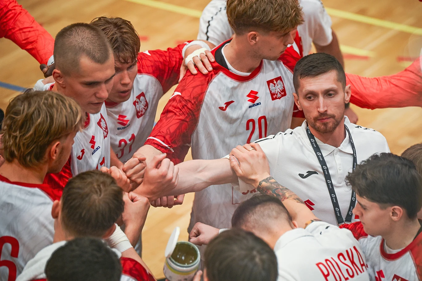 Grupa polskich zawodników w czerwono-białych strojach sportowych tworzy zwarty krąg, łącząc dłonie na znak jedności i wspólnego celu, przy jednym z nich stojący trener w białej koszulce.