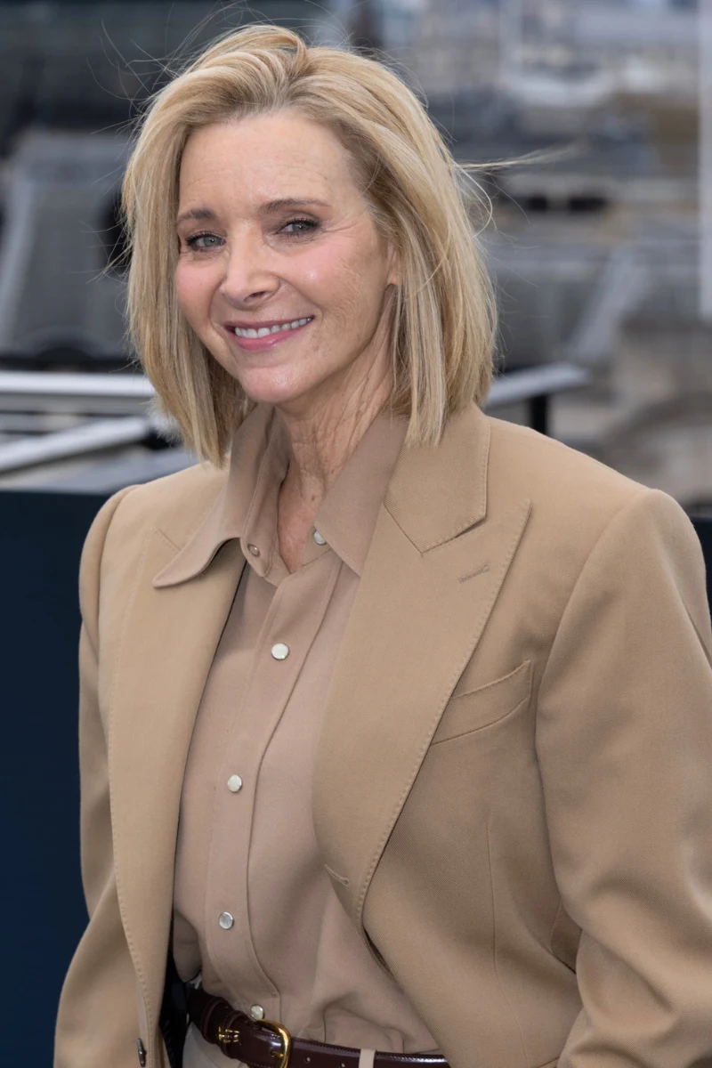 Lisa Kudrow kończy 63 lata Kobieta o blond włosach w eleganckiej, beżowej marynarce i koszuli, delikatnie uśmiechnięta, stoi na tle zabudowań miejskich.