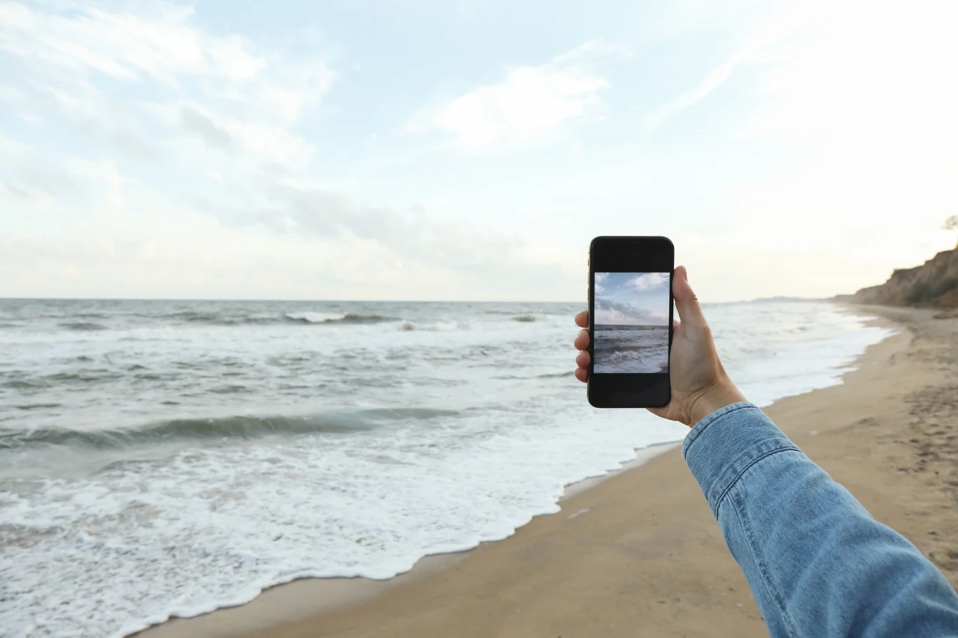 Ręka w niebieskiej kurtce trzyma smartfon, na którego ekranie widać wybrzeże z falującym morzem i jasnym niebem, tło stanowi piaszczysta plaża oraz rozległy horyzont.