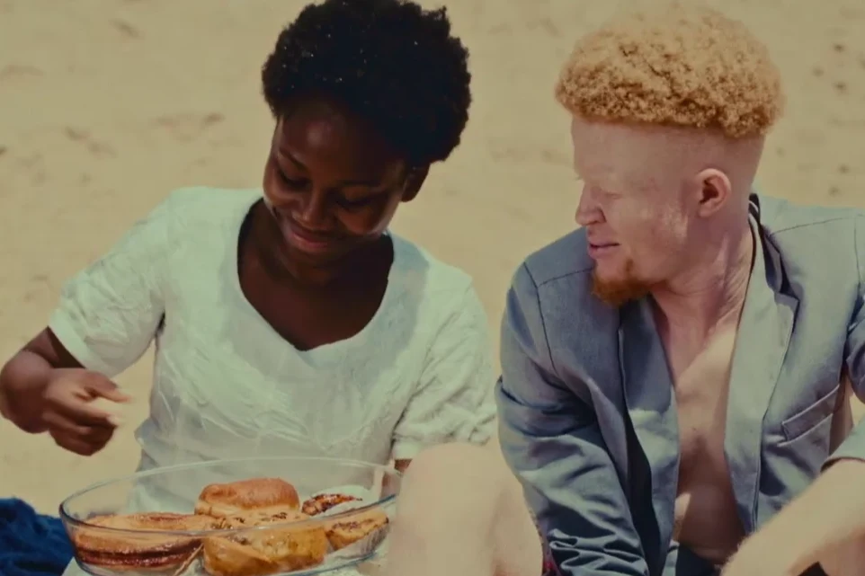 Kadr ze zwiastunu filmu "Moja Semba" Kadr z filmu „Moja Semba”: dwie osoby siedzą na plaży, jedna z nich podaje jedzenie z dużej miski.
