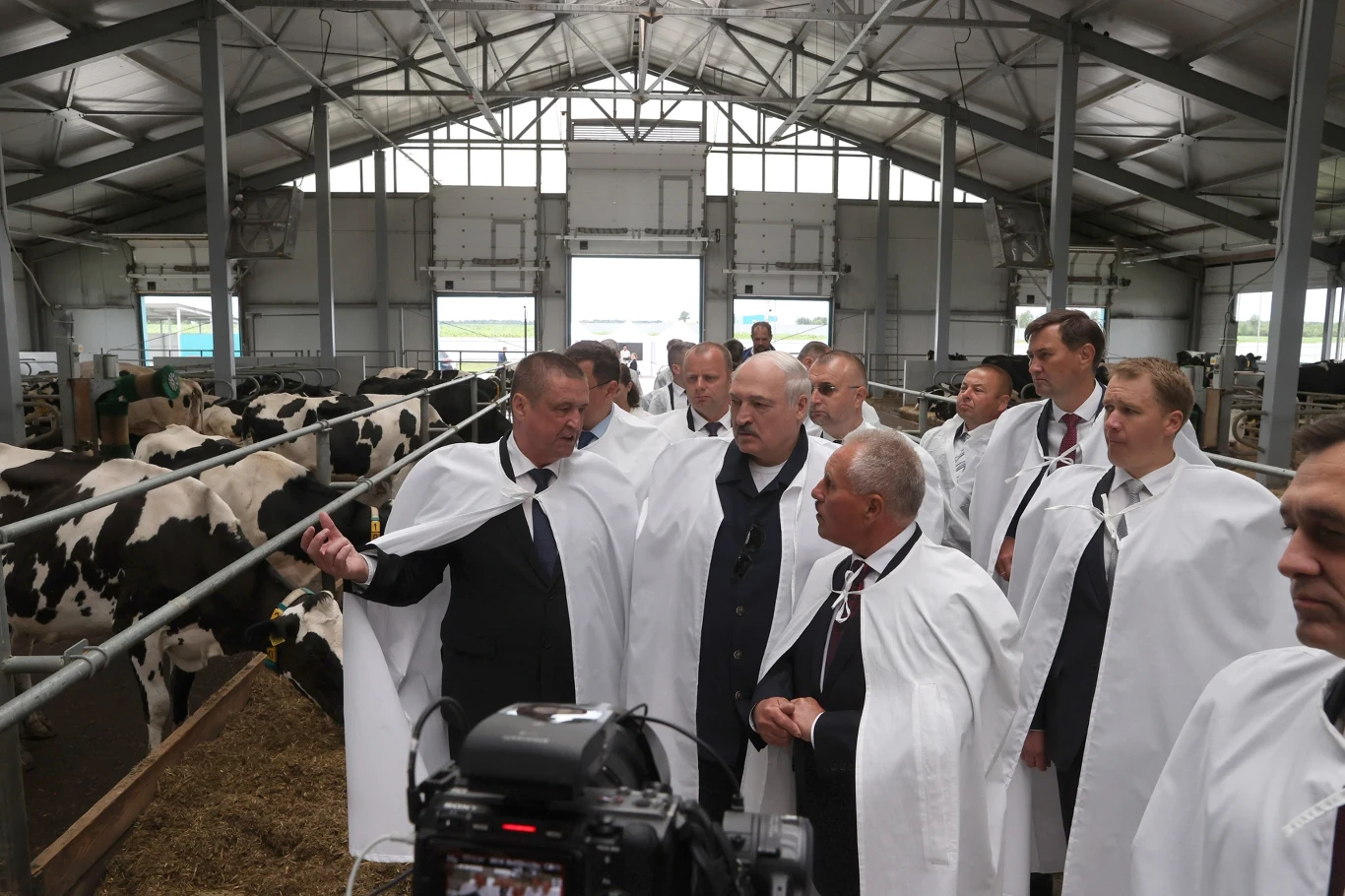 Grupa mężczyzn ubranych w białe fartuchy rozmawia w nowoczesnej oborze, pośród krów stojących w rzędach przy wygrodzeniu.