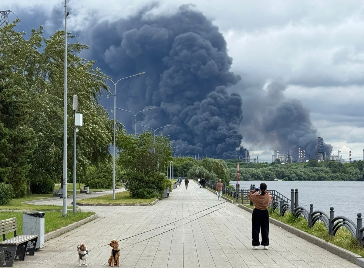 Gęsty, czarny dym unoszący się nad dużym przemysłowym kompleksem po drugiej stronie rzeki, na pierwszym planie ludzie fotografujący zdarzenie oraz osoba z dwoma psami na smyczy na szerokiej promenadzie.