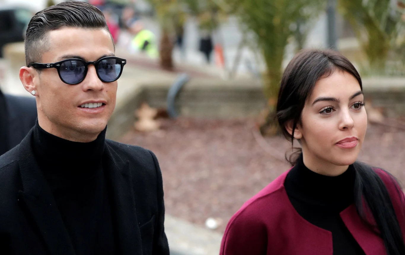 Cristiano Ronaldo i Georgina Rodriguez Dwójka elegancko ubranych osób idących razem na tle miejskiej scenerii, mężczyzna w ciemnych okularach i czarnym golfie, kobieta w bordowej marynarce z czarnym topem.