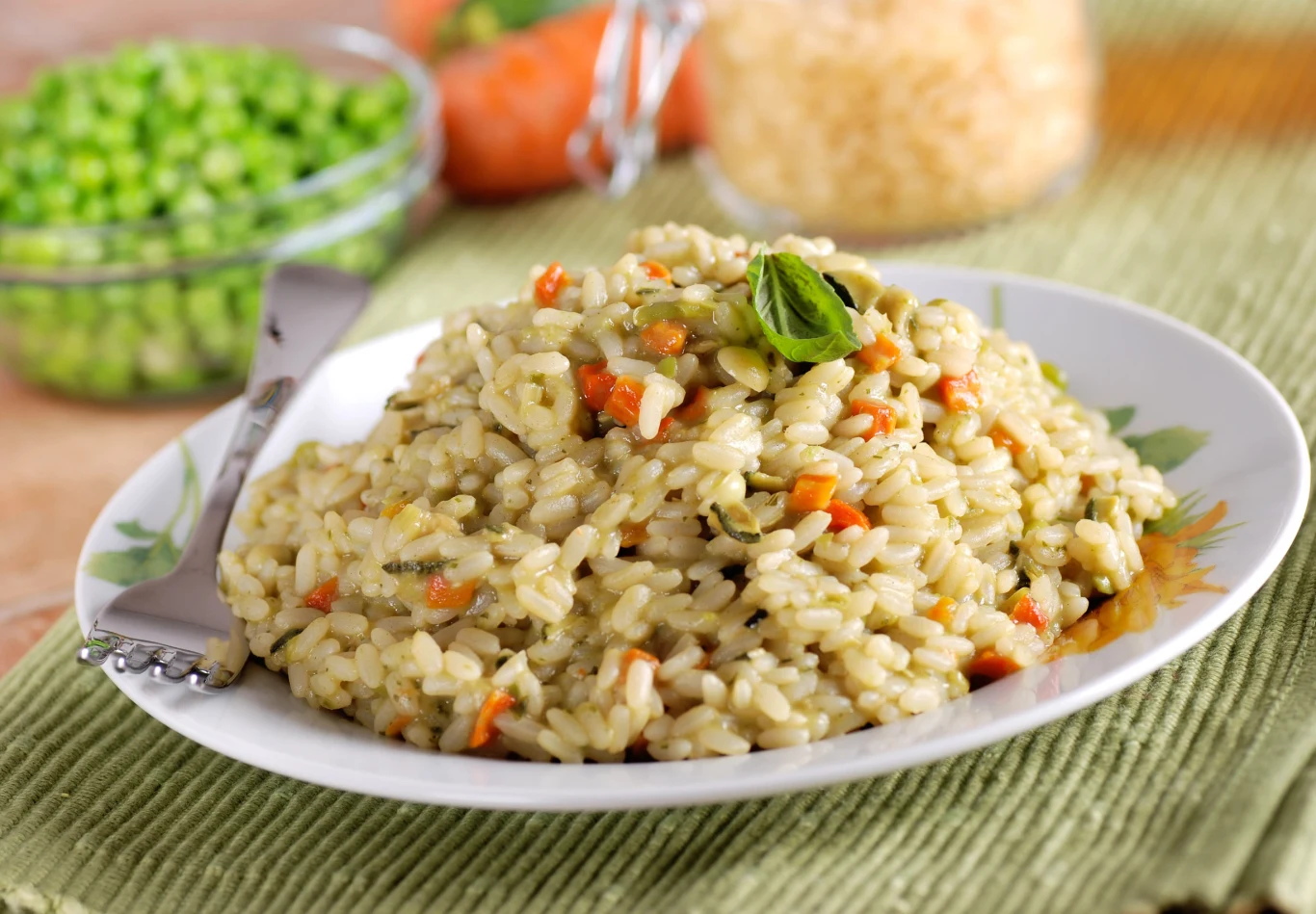 Włoskie risotto zrobisz w domu. Będzie tak pyszne, jak w restauracji Porcja kremowego risotto z marchewką, groszkiem i ziołami, udekorowana bazylią na białym talerzu.