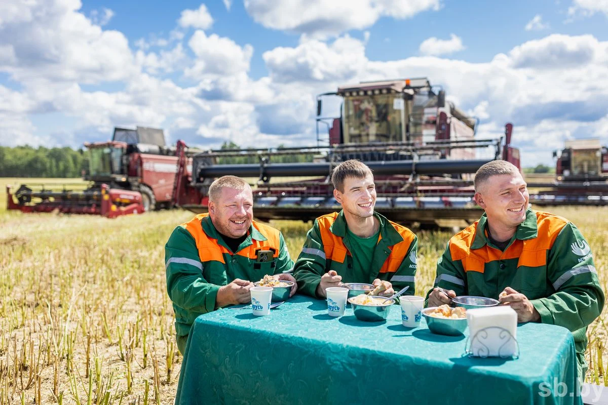 Trzech mężczyzn w zielono-pomarańczowych kombinezonach siedzi przy stole na polu, jedząc posiłek i śmiejąc się, w tle widoczne są kombajny zbożowe i błękitne niebo z chmurami.