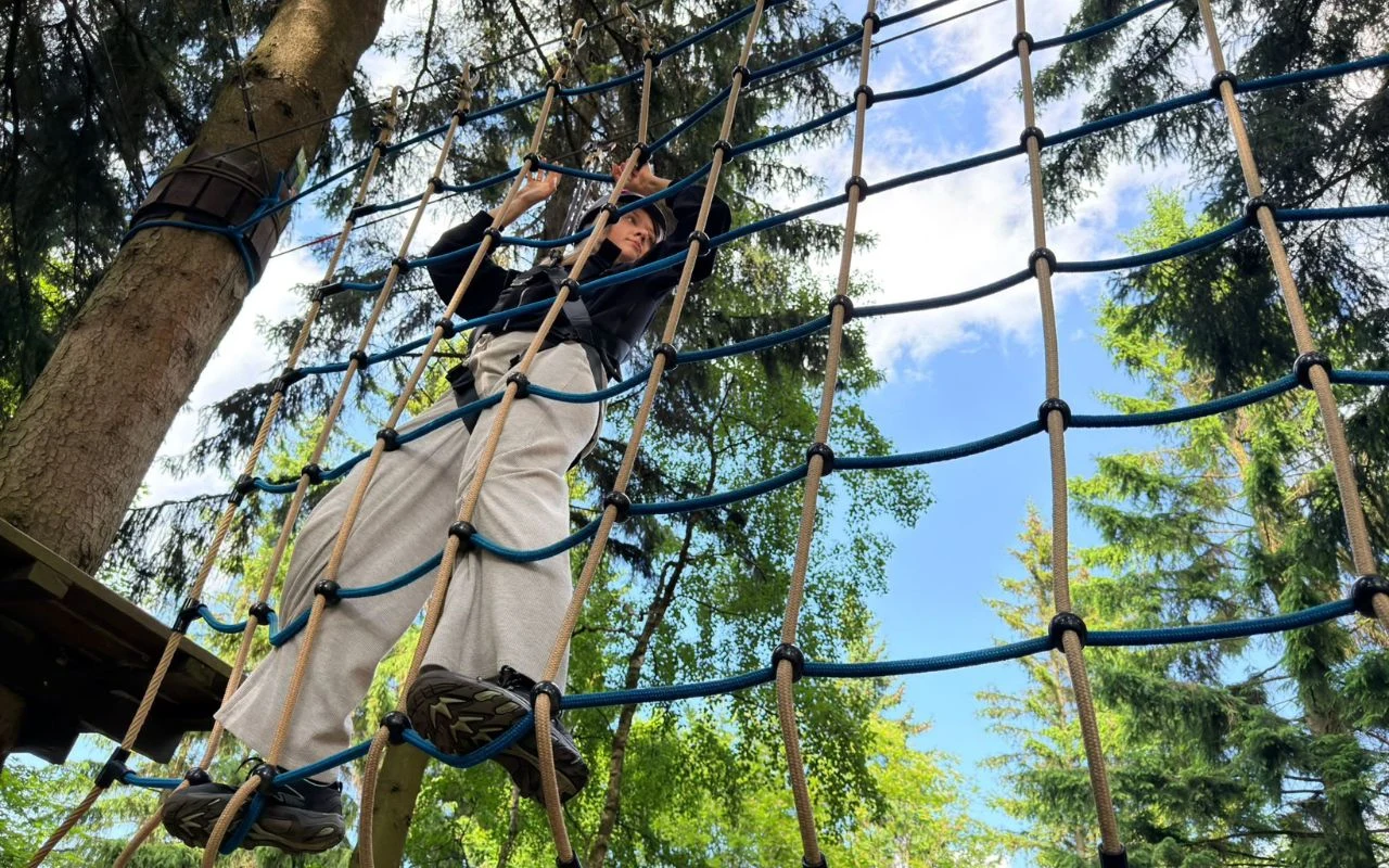Osoba ubrana na sportowo wspinająca się po linowej siatce umieszczonej między drzewami w parku linowym, otoczona zielonymi koronami drzew i niebem w słoneczny dzień.