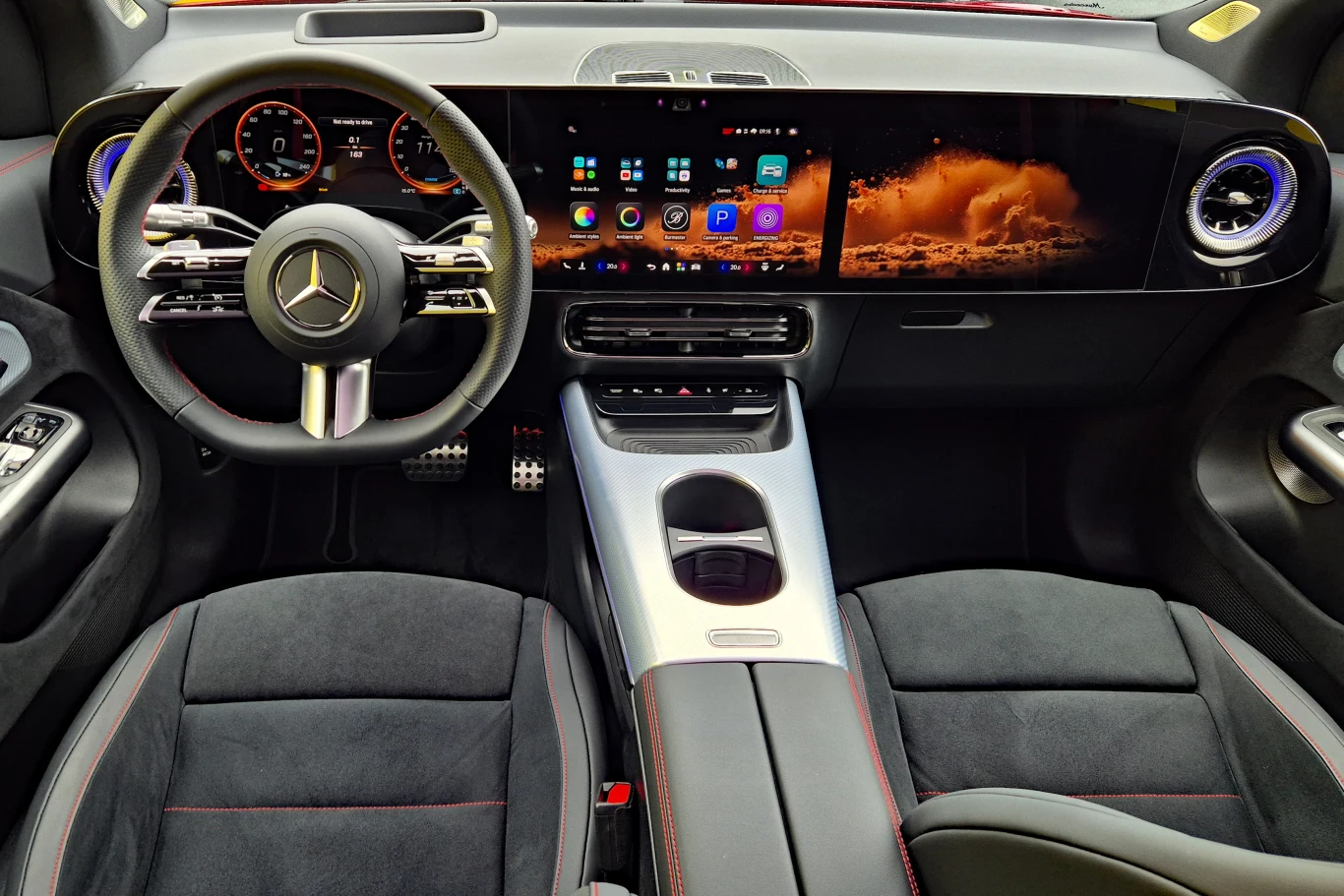 Kokpit luksusowego samochodu marki Mercedes-Benz z nowoczesną kierownicą, elektronicznymi zegarami, dużym panoramicznym ekranem dotykowym i elegancką konsolą środkową wykończoną srebrnym panelem.