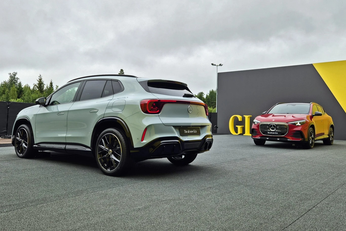 Dwa nowoczesne SUV-y zaparkowane obok siebie na tle ciemnoszarej ściany z żółtym akcentem i napisem „GLA”, jeden w kolorze białym, drugi czerwonym.
