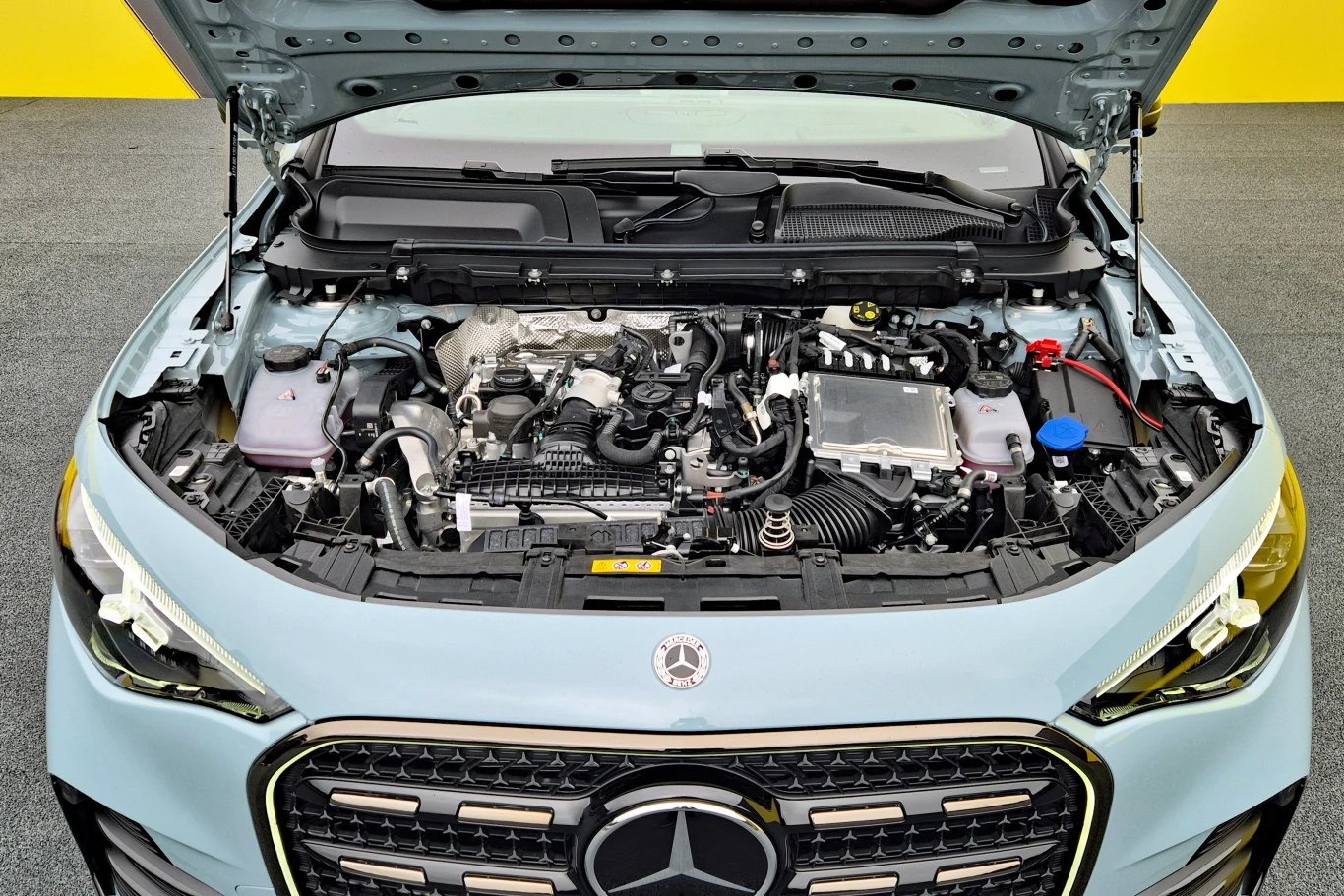 Silnik samochodu marki Mercedes widoczny po podniesieniu maski, z wyraźnym logo na atrapie chłodnicy i elementami technicznymi układu napędowego oraz płynami eksploatacyjnymi.