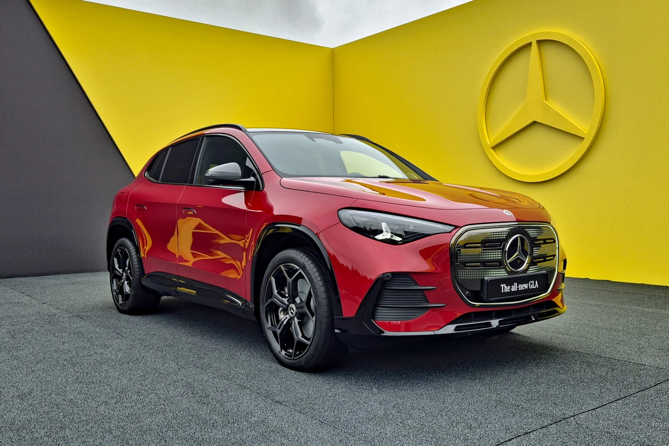 Czerwony samochód marki Mercedes-Benz ustawiony pod żółtą ścianą z dużym logo producenta, nowoczesna sylwetka SUV-a oraz kontrastująca kolorystyka sceny.