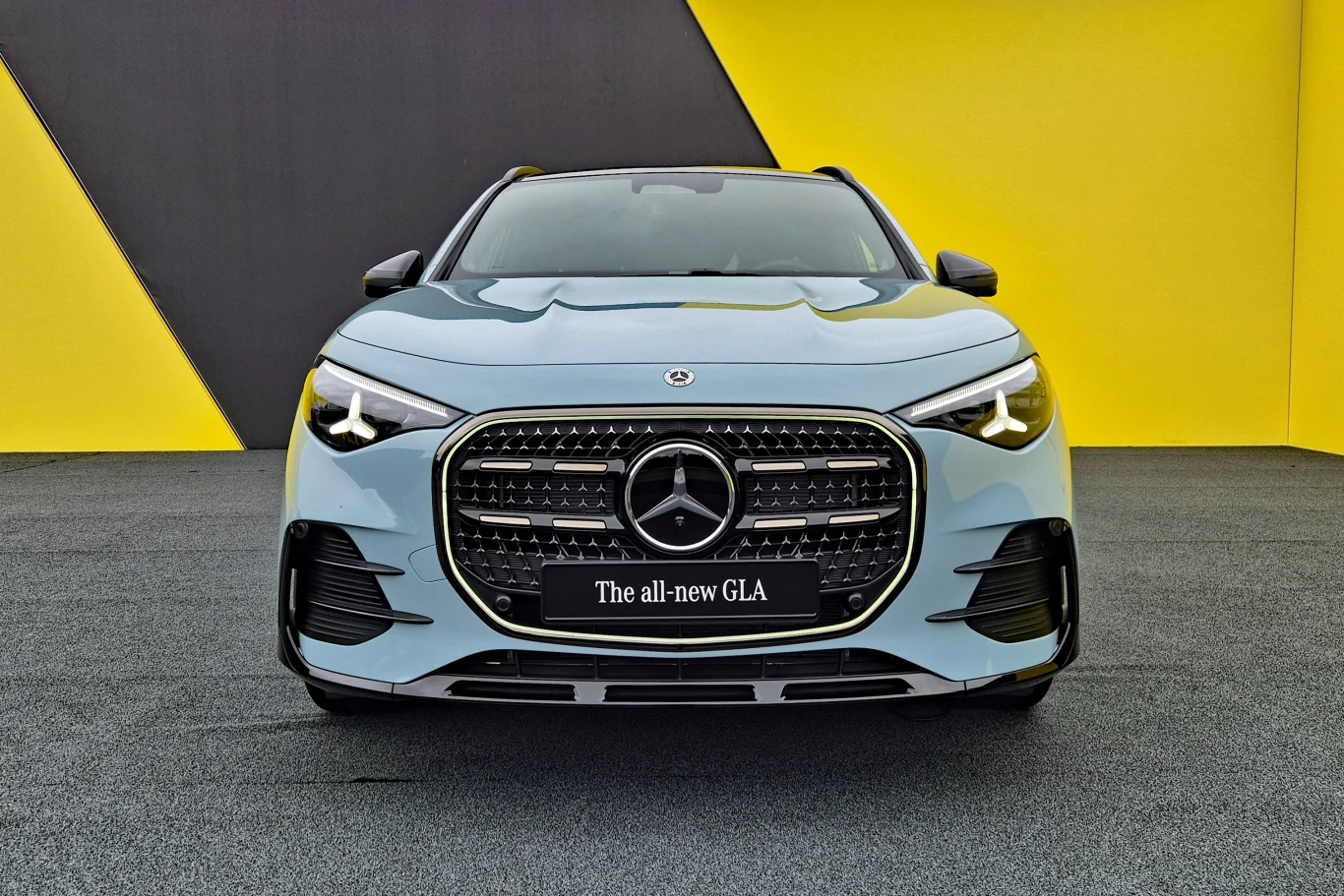 Nowoczesny, jasnoniebieski Mercedes GLA z charakterystycznym grillem i logo marki na tle żółto-czarnej, geometrycznej ściany.