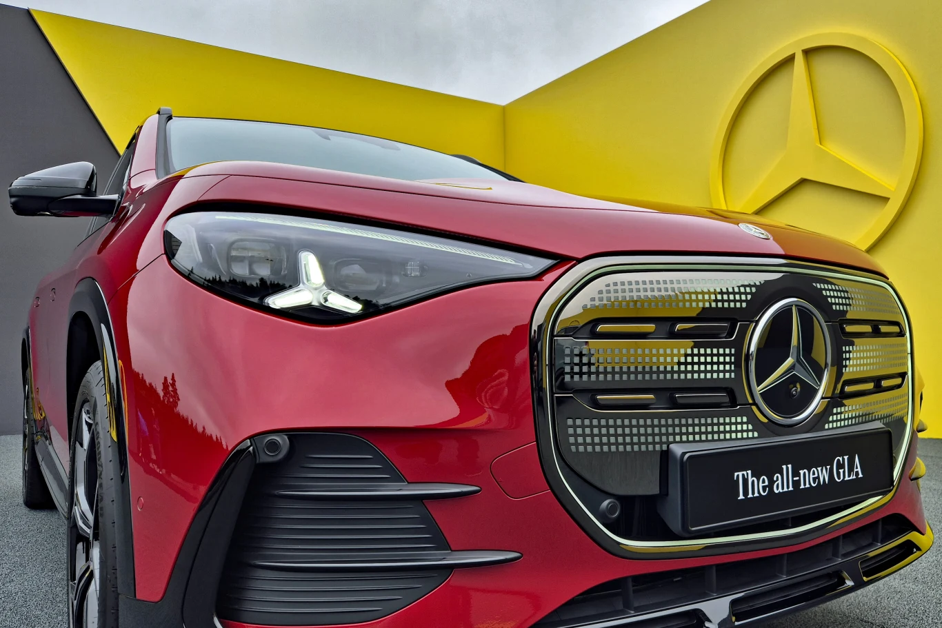 Czerwony Mercedes GLA najnowszej generacji zaparkowany przed geometryczną, żółto-szarą ścianką z charakterystycznym logotypem marki.