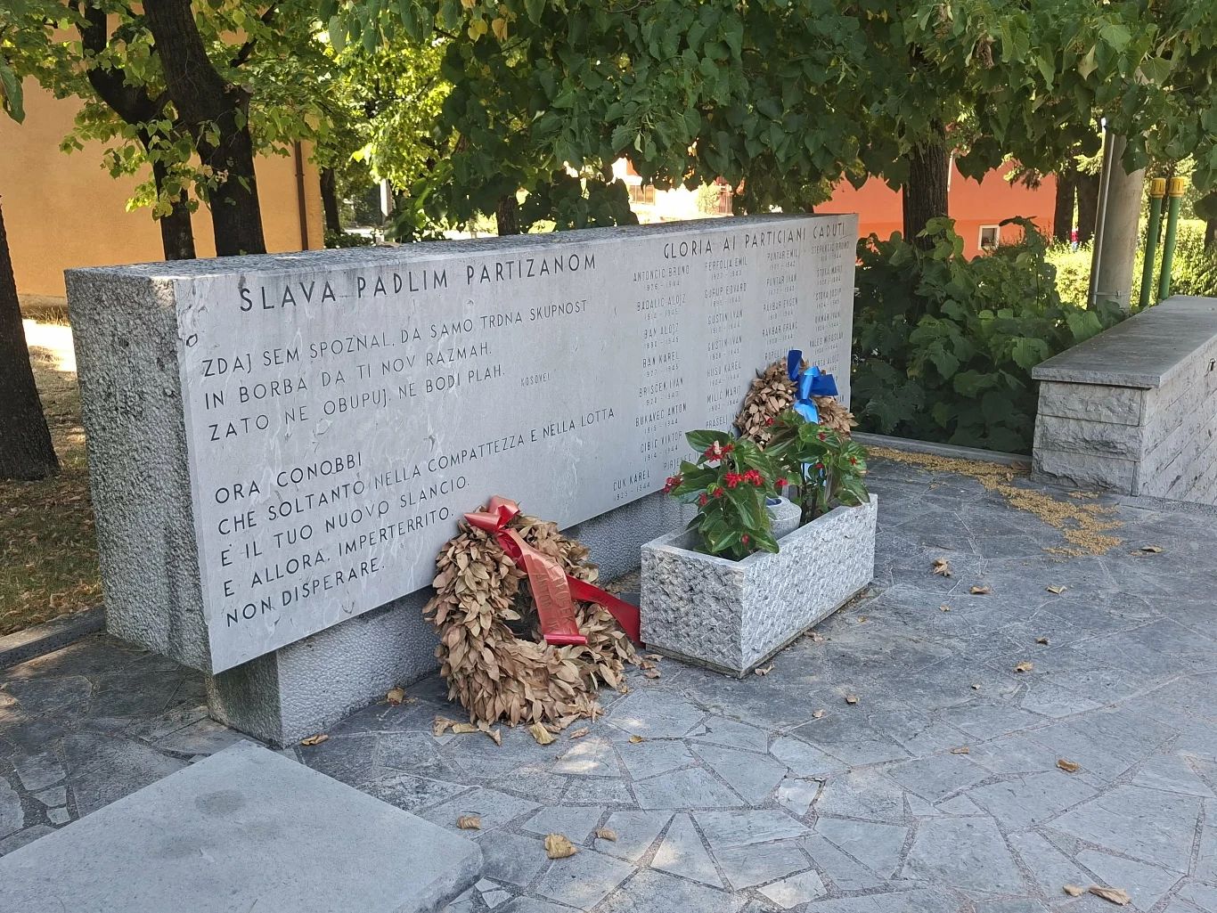 Kamienny pomnik z wyrytym napisem upamiętniającym poległych partyzantów, ozdobiony dwoma wieńcami i donicą z kwiatami, ustawiony pod rozłożystym drzewem na wybrukowanym placu.
