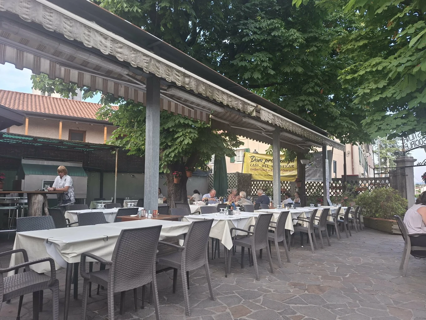 Zewnętrzny taras restauracji z dużym stołem nakrytym białym obrusem, otoczony krzesłami, osłonięty markizą i otoczony zielonymi drzewami, kilka osób siedzi i rozmawia, a kelner podaje dania.
