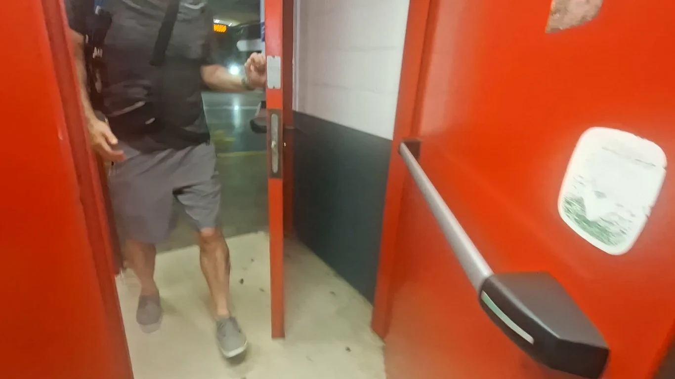 Wejście jak do magazynu, a nie w stronę autobusowych stanowisk Mężczyzna w szarych szortach i szarych butach przechodzi przez czerwone drzwi, w tle fragment parkingu podziemnego.