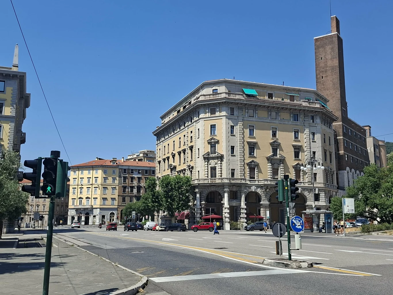 Dzień dobry w Trieście Monumentalny narożny budynek z kolumnami i arkadami przy szerokiej, pustej ulicy, otoczony kilkoma drzewami i sąsiadującymi kamienicami; po prawej wysoka, ceglana wieża dominująca nad okolicą.