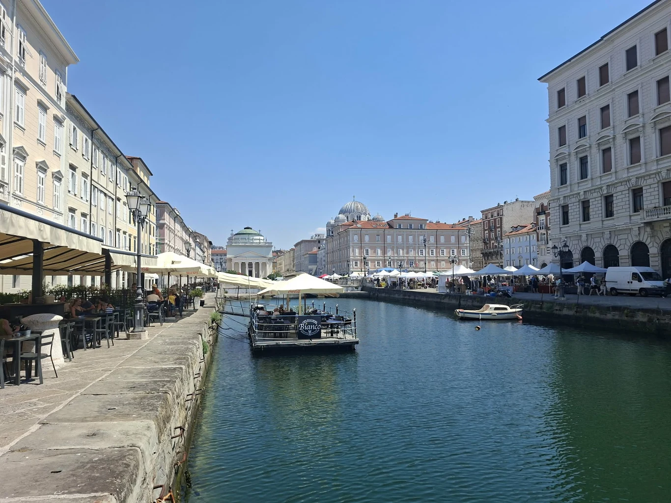 Canale Grande w Trieście Kanał otoczony klasycznymi kamienicami, z ustawionymi wzdłuż nabrzeża stolikami restauracyjnymi pod białymi parasolami oraz zacumowanymi łodziami na spokojnej wodzie, w tle widoczna monumentalna kopuła kościoła.