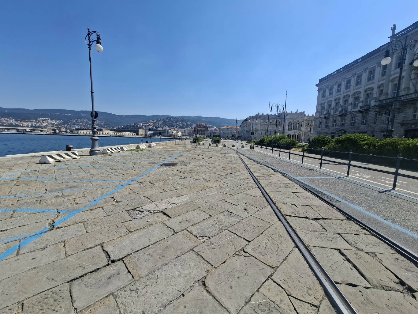 Bulwar przy porcie w Trieście Kamienna promenada z biegnącymi torami tramwajowymi, latarnią uliczną oraz historycznymi budynkami po prawej stronie, z widokiem na zatokę i miasto w oddali.