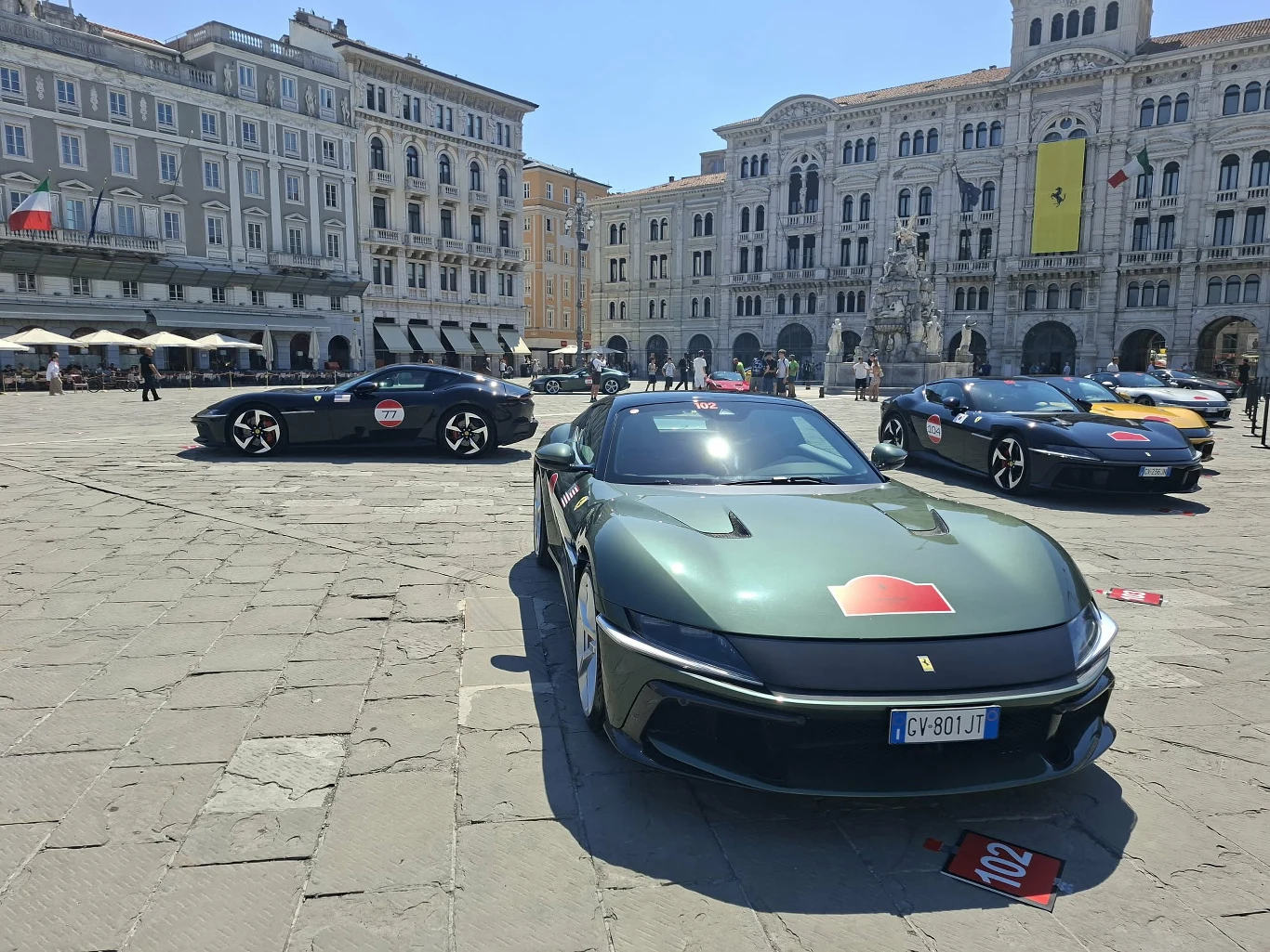 Egzemplarze Ferrari na Piazza Unità d'Italia Trzy sportowe samochody Ferrari ustawione na brukowanym placu otoczonym zabytkowymi kamienicami i reprezentacyjnym budynkiem z flagą Ferrari na fasadzie.