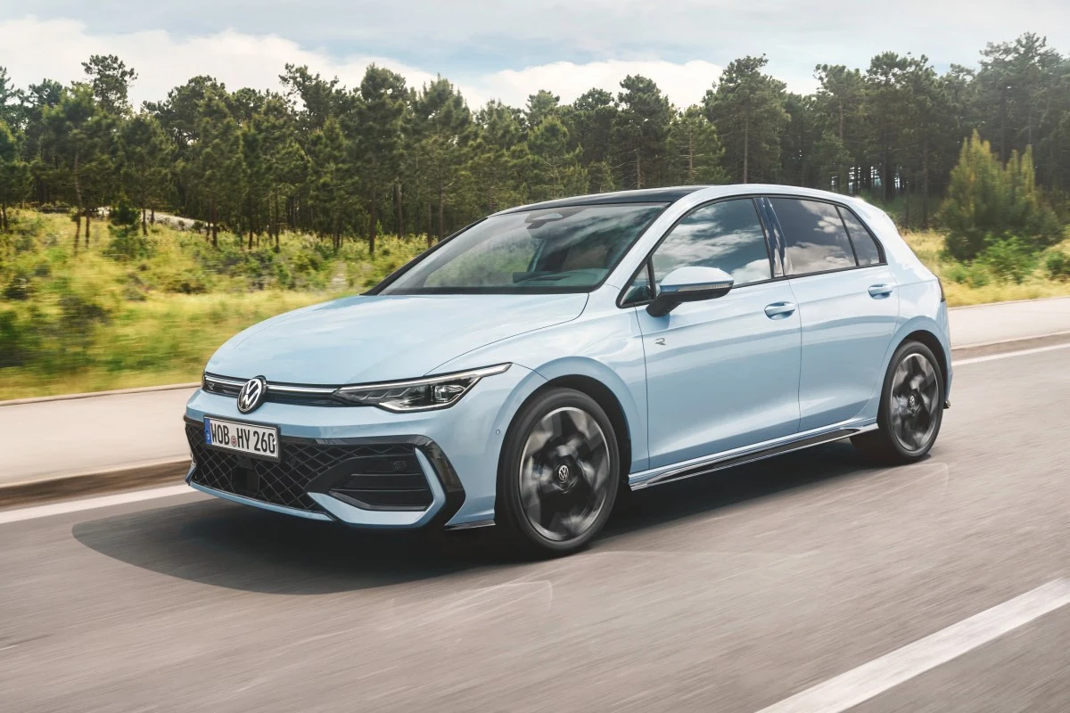 Jasnoniebieski Volkswagen Golf ósmej generacji w wersji hatchback porusza się po asfaltowej drodze, otoczonej zielenią lasu.