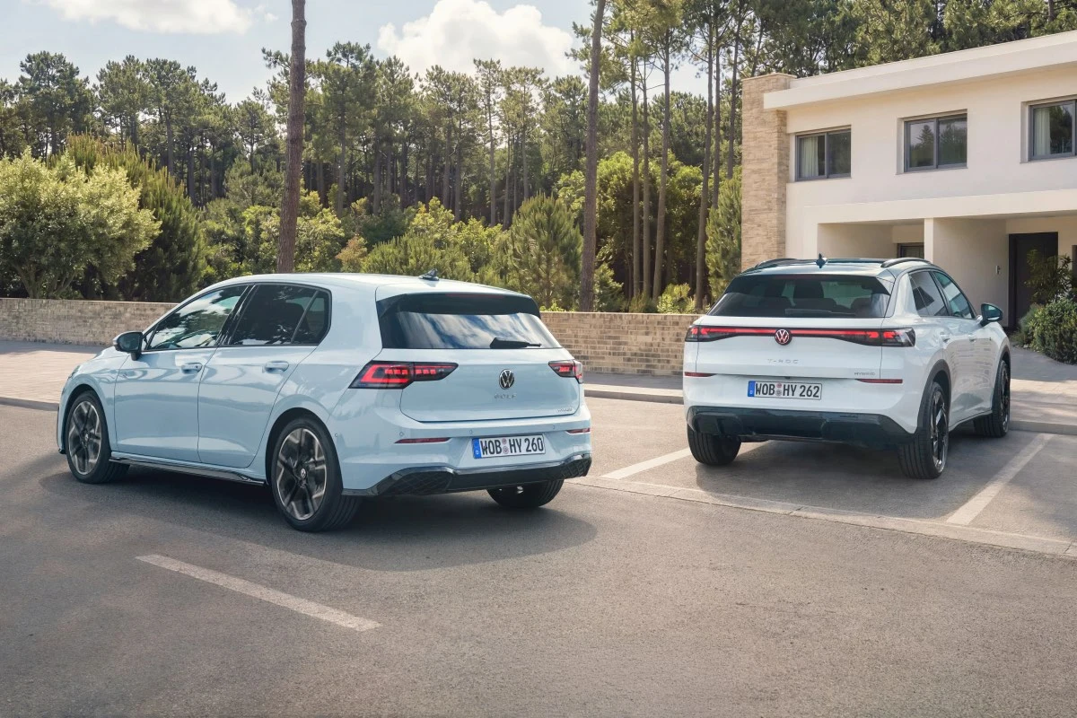 Dwa nowoczesne samochody marki Volkswagen zaparkowane obok siebie na pustym placu, na tle jasnego budynku oraz lasu sosnowego w słoneczny dzień.