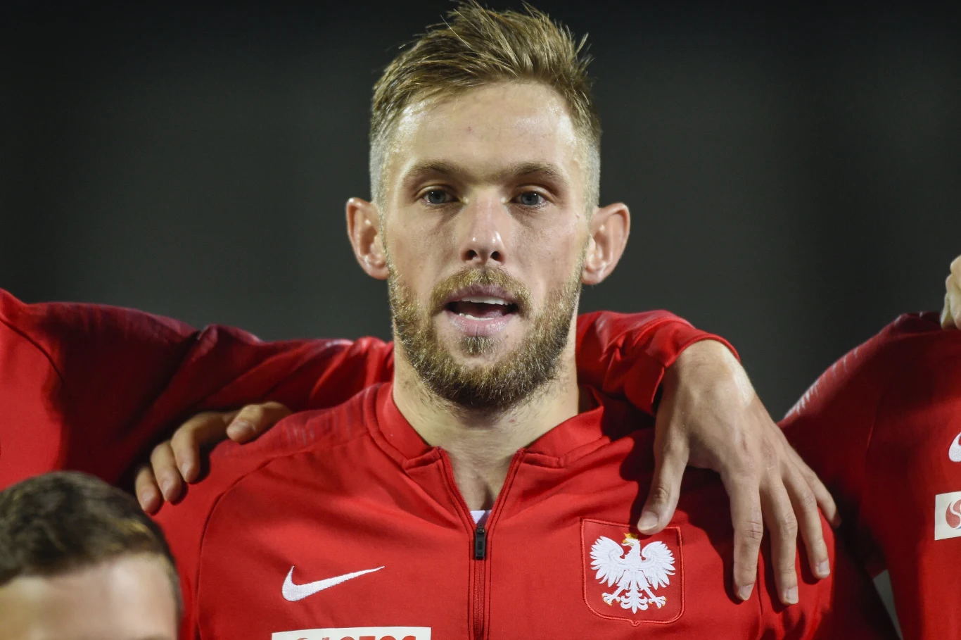 Maciej Rybus znów zagra w Polsce Piłkarz w czerwonym sportowym dresie z godłem Polski na piersi, stojący w szeregu z drużyną, z ramionami kolegów opartymi na jego barkach.