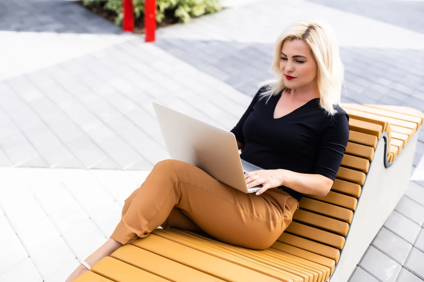 Kobieta o blond włosach w czarnej bluzce i brązowych spodniach pracuje na laptopie, siedząc na nowoczesnej drewnianej ławce na świeżym powietrzu.