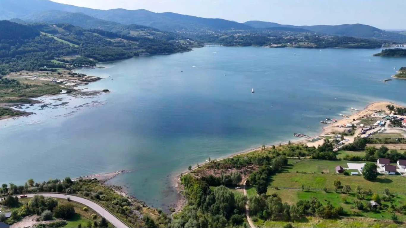 Jezioro Mucharskie Rozległe jezioro otoczone zielonymi wzgórzami oraz plażą z kilkoma budynkami, w tle widoczne są góry.