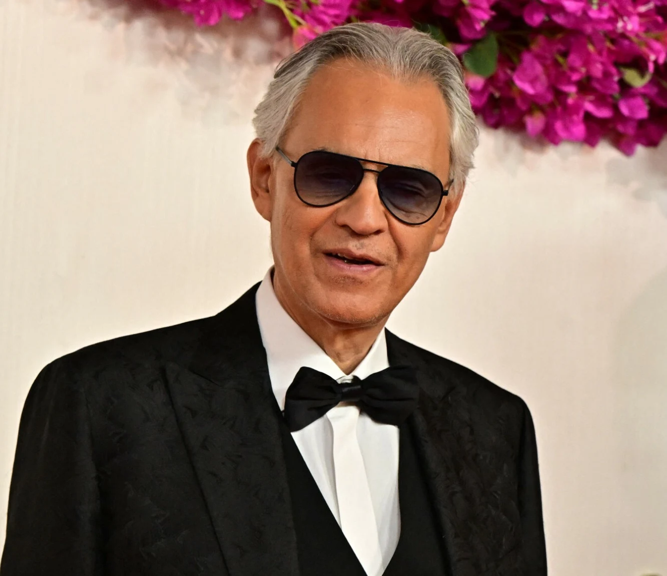 Andrea Bocelli Elegancki mężczyzna w ciemnym garniturze i muchą, w okularach przeciwsłonecznych, na tle jasnej ściany ozdobionej różowymi kwiatami.