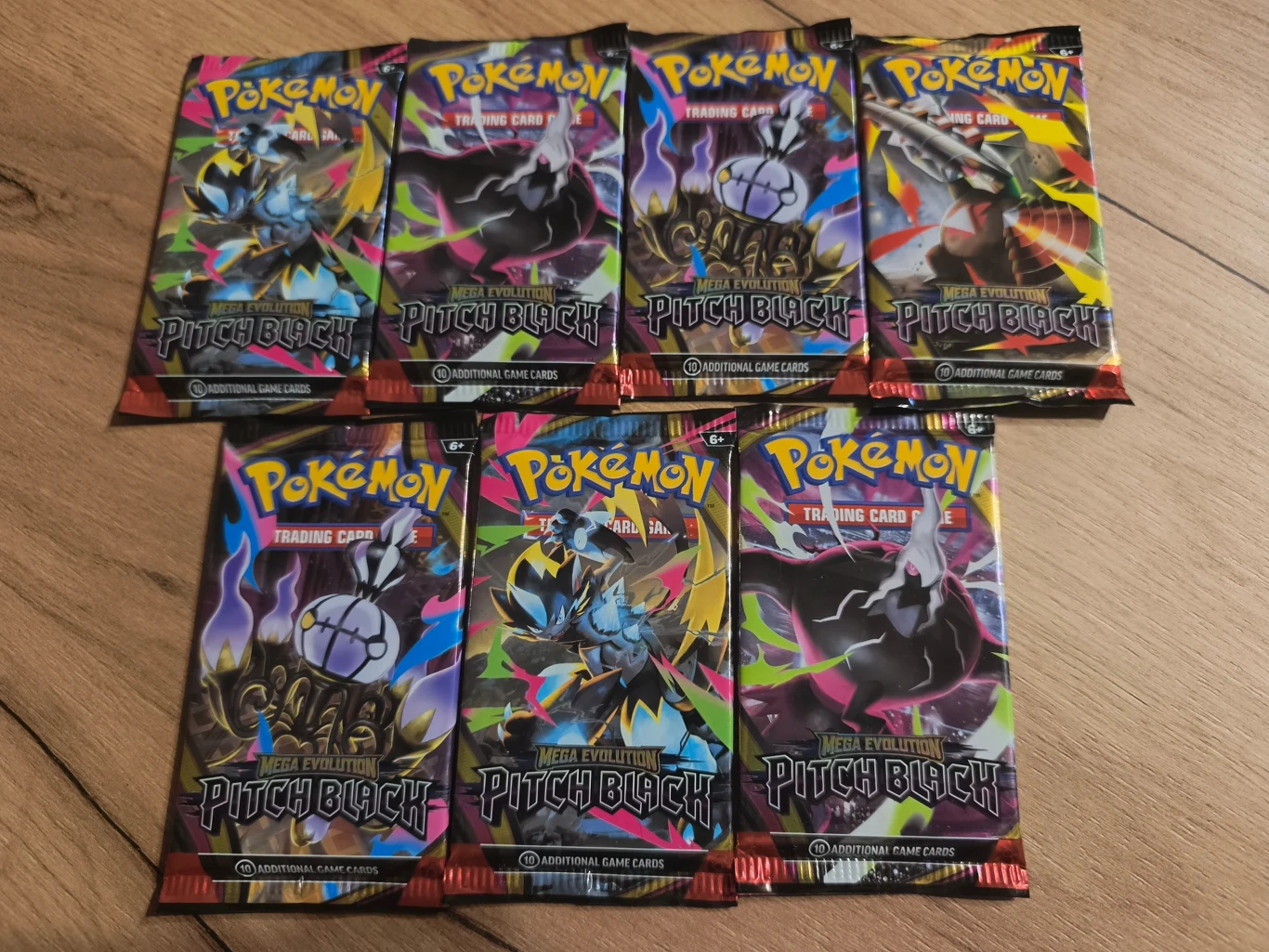 Siedem zamkniętych boosterów kart Pokémon Pitch Black ułożonych na drewnianej podłodze, każdy z wyrazistą grafiką przedstawiającą różne postacie Pokémonów na ciemnym tle.