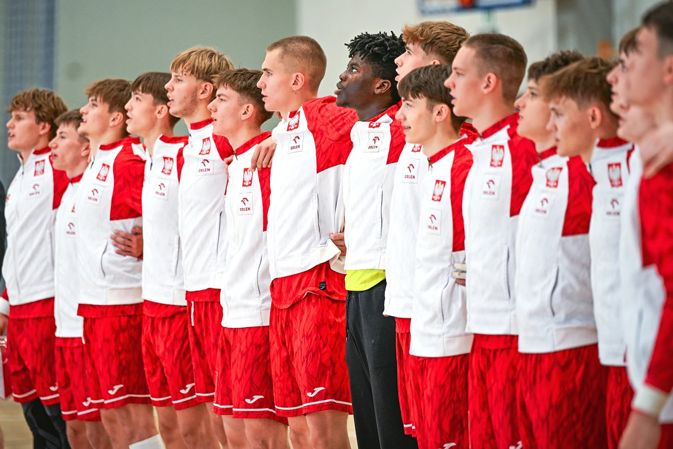 Grupa młodych sportowców w biało-czerwonych dresach z godłem Polski na piersi stoi ramię w ramię, tworząc jednolitą drużynę gotową do rywalizacji.